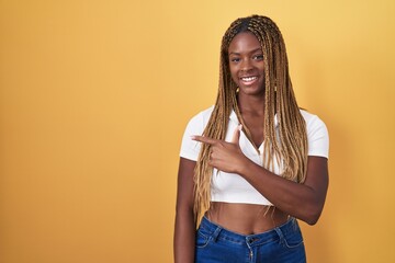 Wall Mural - African american woman with braided hair standing over yellow background cheerful with a smile on face pointing with hand and finger up to the side with happy and natural expression