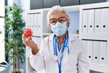 Wall Mural - Senior grey-haired woman wearing doctor uniform and medical mask holding virus toy at clinic