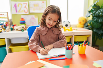adorable hispanic girl preschool student sitting on table writing on notebook at kindergarten