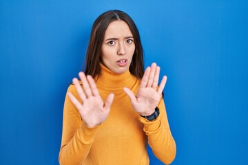 Poster - Young brunette woman standing over blue background moving away hands palms showing refusal and denial with afraid and disgusting expression. stop and forbidden.