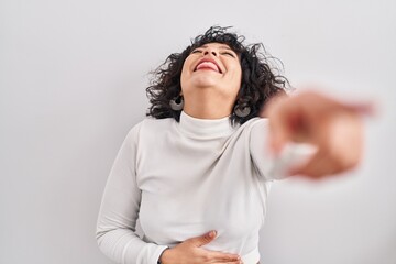 Hispanic woman with curly hair standing over isolated background laughing at you, pointing finger to the camera with hand over body, shame expression