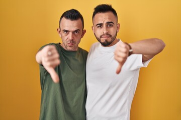 Sticker - Homosexual couple standing over yellow background looking unhappy and angry showing rejection and negative with thumbs down gesture. bad expression.