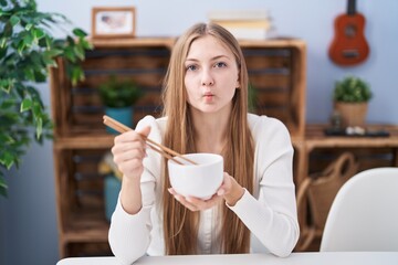 Canvas Print - Young caucasian woman eating asian food using chopsticks making fish face with mouth and squinting eyes, crazy and comical.