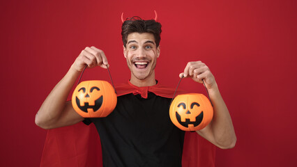 Wall Mural - Young hispanic man wearing devil costume holding halloween pumpkin baskets over isolated red background