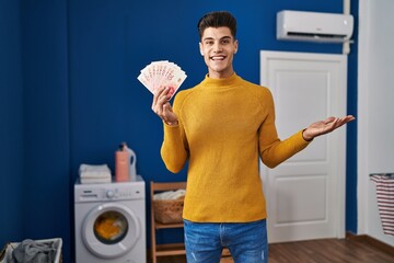 Wall Mural - Young hispanic man at laundry room holding shekels celebrating achievement with happy smile and winner expression with raised hand