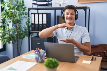 Wall Mural - Young hispanic man working at the office wearing headphones gesturing with hands showing big and large size sign, measure symbol. smiling looking at the camera. measuring concept.