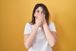 Young brunette woman standing over yellow background shocked covering mouth with hands for mistake. secret concept.