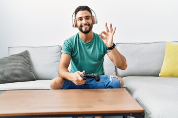 Wall Mural - Handsome hispanic man wearing headphones playing video game holding controller doing ok sign with fingers, smiling friendly gesturing excellent symbol