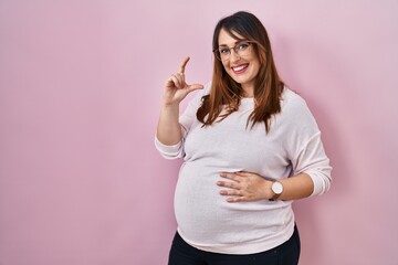 Poster - Pregnant woman standing over pink background smiling and confident gesturing with hand doing small size sign with fingers looking and the camera. measure concept.