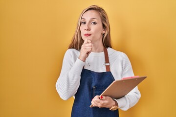 Poster - Young blonde woman wearing professional waitress apron holding clipboard serious face thinking about question with hand on chin, thoughtful about confusing idea