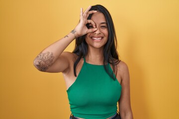 Canvas Print - Brunette woman standing over yellow background doing ok gesture with hand smiling, eye looking through fingers with happy face.