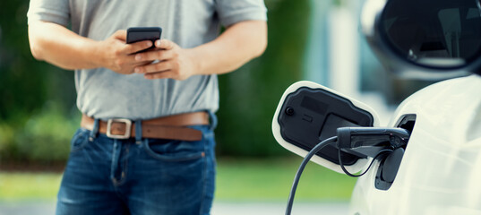 Sticker - Progressive asian man install cable plug to his electric car with home charging station in the backyard. Concept use of electric vehicles in a progressive lifestyle contributes to clean environment.