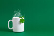Saint Patricks Day background. White cup of hot tea or coffee with steam for St. Patrick's day, mug, green paper clover leaf on green background. Front view