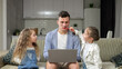 Junior schoolgirl paints father lips and enjoys playing with sister. Father with exhausted expression tries to work on laptop getting distracted at home