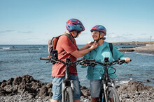 Happy Caucasian Senior Couple Wearing Helmets Riding On The Beach With Electric Bicycles. Authentic Elderly Retired Life Concept. Horizon Over Sea