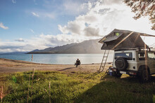 A Woman Sits Along A Lake Next To A SUV With A Tent In New Zealand