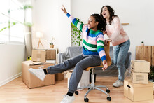 Carefree Young Woman Pushing Friend Sitting In Swivel Chair At New Home