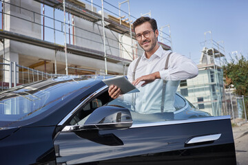 Smiling architect with tablet PC standing by car at construction site