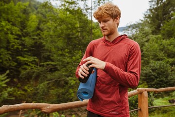 Wall Mural - Young white man holding mat and thermos bottle during workout in forest