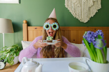 Cheerful Woman Wearing Sunflower Sunglasses Holding Plate With Cupcake In Bed At Home