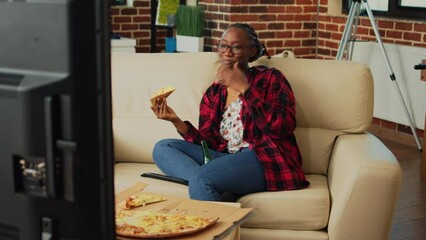 Wall Mural - Cheerful woman eating slice of pizza and watching film, having fun with favorite tv show and food delivery meal. Young person enjoying takeout order and bottles of beer, watch movie.