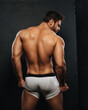 Naked muscled man in white boxer underwear on black background. Sexy back of bodybuilder in studio. Backside of muscular male model holding his underwear.