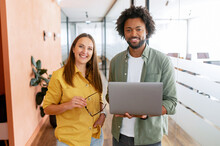 Two Happy Diverse Colleagues African American Man And Caucasian Woman Looking At Camera Standing In Office Lobby Hall, The Man Is Holding Laptop