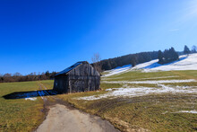 An Old Dilapidated Wooden Hut In February On A Sunny Day With Blue Sky In A Meadow Partly Covered With Snow