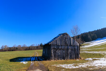An Old Dilapidated Wooden Hut In February On A Sunny Day With Blue Sky In A Meadow Partly Covered With Snow