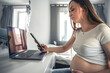 © puhimec - Young pregnant woman uses smartphone and laptop at home.