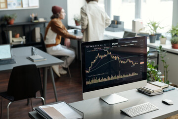 Fototapete - Workplace of financial analyst or trader with graphic data on screen of computer monitor standing on desk against two colleagues