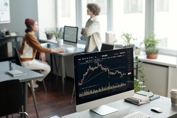 Fototapete - Computer monitor with graphic financial statistics on screen standing on workplace of broker or trader against two young economists