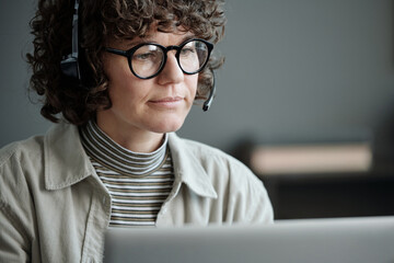 Aufkleber - Young serious brunette female operator of call center in headset and eyeglasses sitting in front of laptop screen and listening to online client