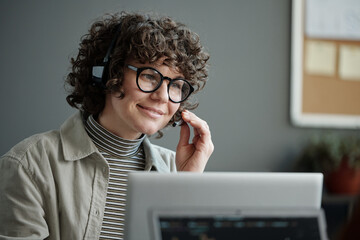 Aufkleber - Young smiling female IT support engineer in headset looking at laptop screen and answering questions of online clients in office