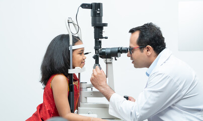 Indian middle aged professional ophthalmologist or optometrist man work at optometry clinic, using optometry instruments little girl patient to check eyesight and eyes vision at modern clinic