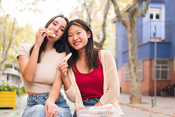 Wall Mural - portrait of a happy female couple sharing some sweet buns sitting in a city park, concept of friendship and love between people of the same sex