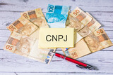 Fototapeta  - A sigla CNPJ referente ao Cadastro Nacional da Pessoa Jurídica em Português do Brasil escrita em um pedaço de papel. Notas do Real Brasileiro sobre uma mesa de madeira.
