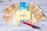 Fototapeta  - A sigla MEI de Microempreendedor Individual escrita em um pedaço de papel. Notas do Real Brasileiro na composição. Economia brasileira e finanças.