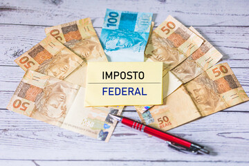 Sticker - A palavra Imposto Federal em Português do Brasil escrita em um pedaço de papel que está sobre notas do Real Brasileiro na composição. Economia brasileira e impostos.