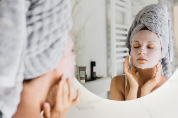 Canvas Print - Woman in bathroom with applied sheet mask on her face looking in the mirror