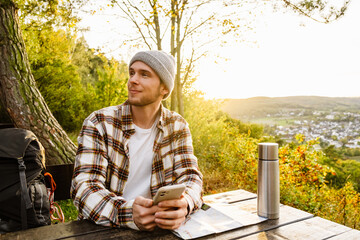 Young man tourist holding mobile phone and looking away while sitting on hillside