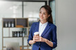 © amnaj - Beautiful Asian business woman standing holding coffee reviewing work and finding new idea to work in the office.