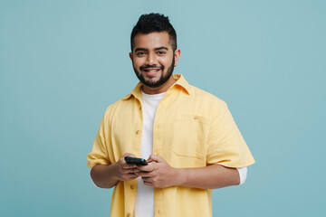 Wall Mural - Smiling indian guy using mobile phone isolated over blue wall