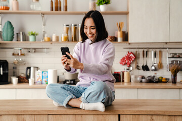 Wall Mural - Cheerful asian woman using mobile phone while sitting on table in kitchen