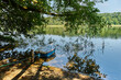 A boat moored in the shade of tree branches on the shore of a lake on a sunny summer morning. The lake is calm and its surface reflects the vegetation growing on its shores.