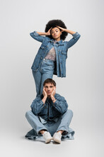 Full Length Of Curly African American Woman Posing Behind Man In Denim Outfit And Sneakers Sitting On Grey.