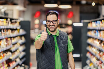 A supermarket manager is standing at marketplace and giving thumbs up while smiling at the camera.