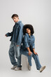 full length of curly african american woman sitting near man in denim clothes on grey.
