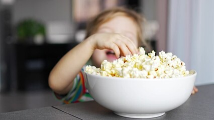 Wall Mural - Little girl is eating popcorn in home kitchen. Focus on hand taking popcorn