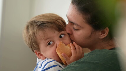 Wall Mural - Affecctionate mother kissing little boy son in cheek while eating bread. Parenting family happiness concept. Mom embracing and hugging child. Candid lifestyle